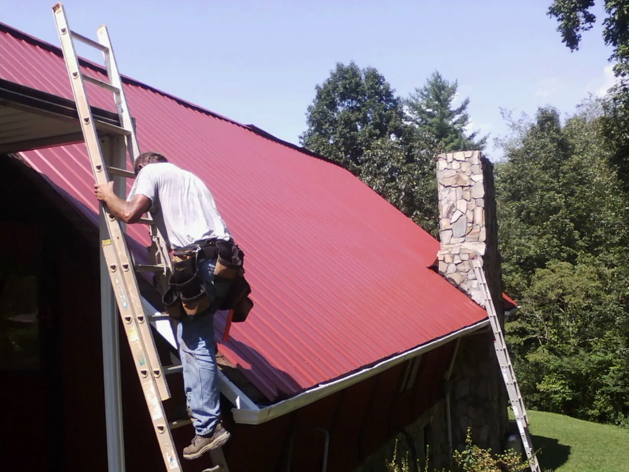 Roofer on a ladder beside a red metal roof and stone chimney — Soffit & Fascia Repair by Roofing Brothers in Burgettstown